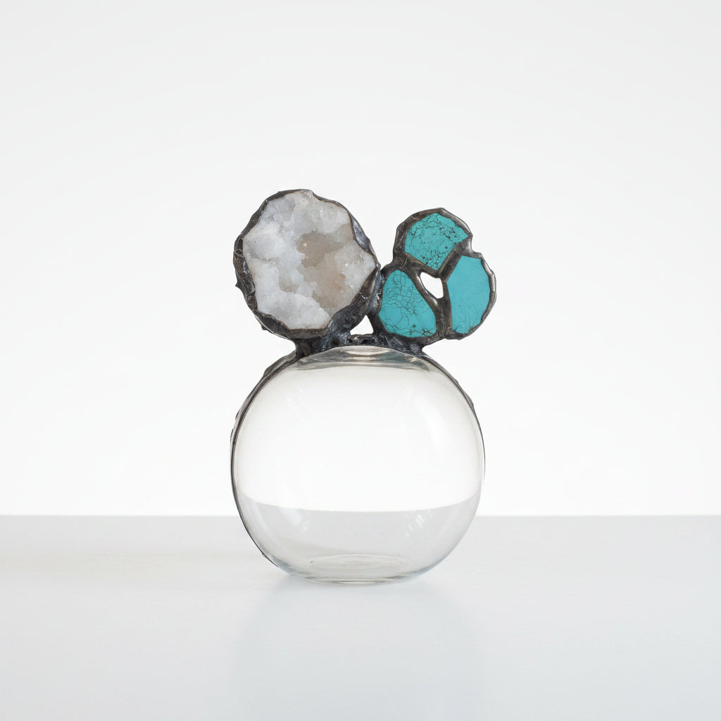 A decorative glass vase with a rounded clear glass body and two sculptural floral or geode-like tops in black, white, and turquoise, set against a light stone wall background.