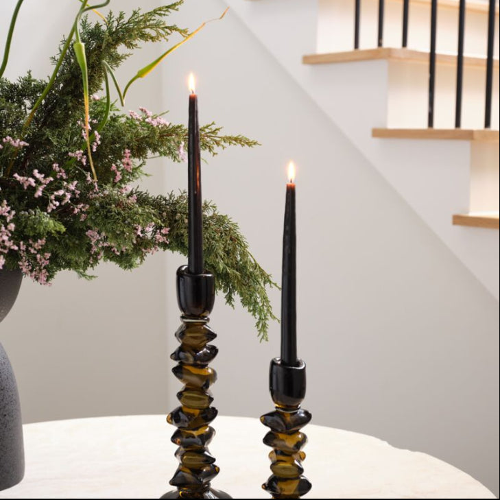 Two black candlesticks with candles on a white surface, with a staircase and plant in the background.