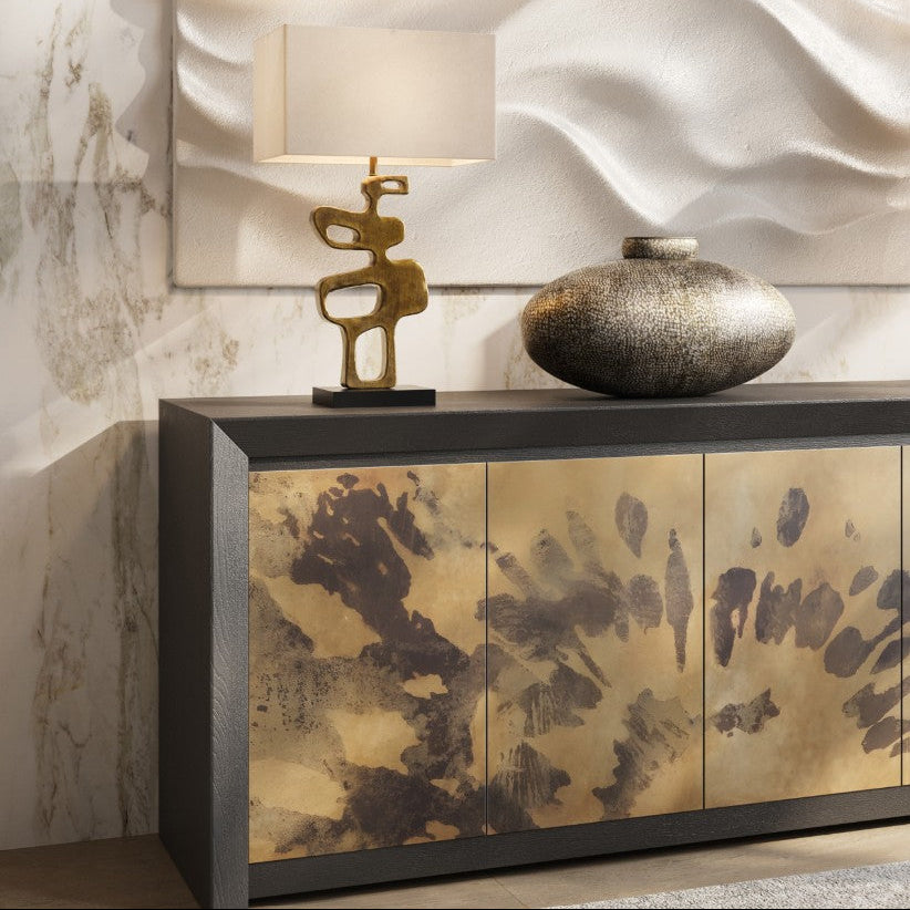 A modern sideboard cabinet with three gold-toned doors featuring dark abstract brushstrokes, topped with a sculptural gold table lamp and a textured metallic vase, set against a textured beige wall.