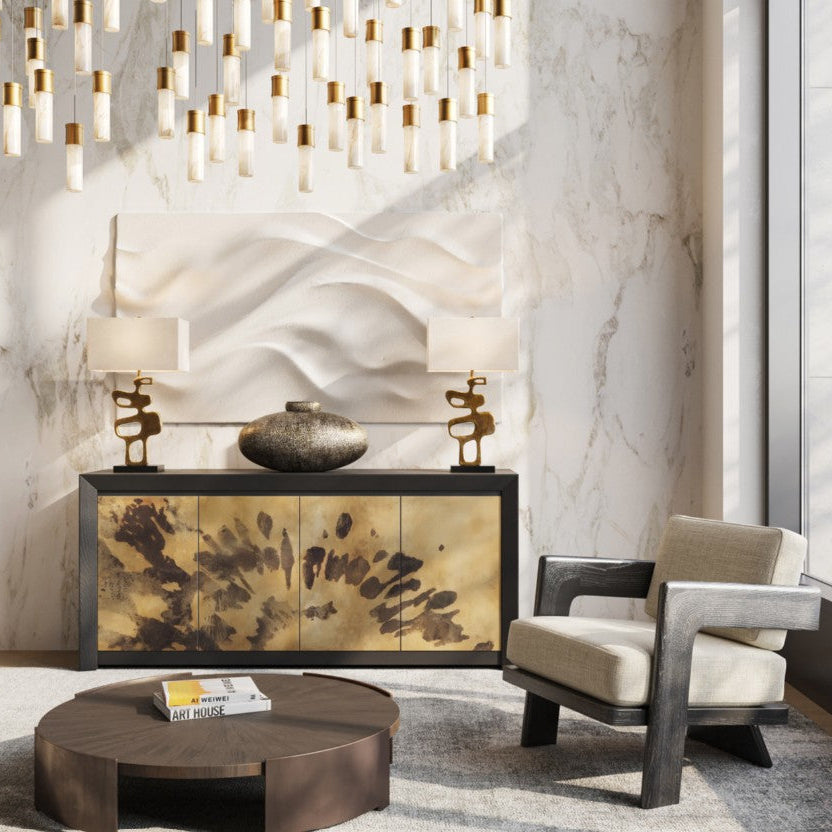 A styled interior scene featuring a modern console cabinet with gold-toned marbled front panels, two sculptural table lamps, a round coffee table, a beige armchair, and a large abstract wall sculpture against a marble-look wall.