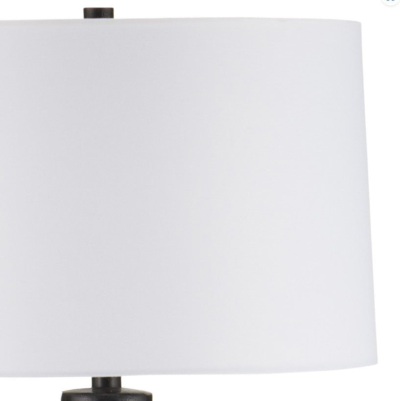 A close-up of a table lamp featuring a large white drum shade and a dark, textured metal base against a plain white background.