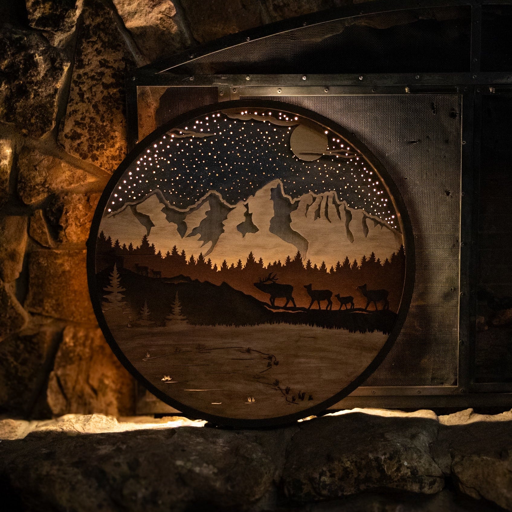 Wooden wall art depicting a night scene with mountains, trees, and animals on a stone surface.