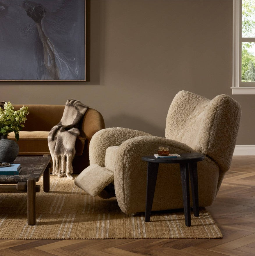 A plush, high-back recliner chair upholstered in a textured beige bouclé or sherpa-like fabric, styled in a warm, modern living room next to a dark wood side table.