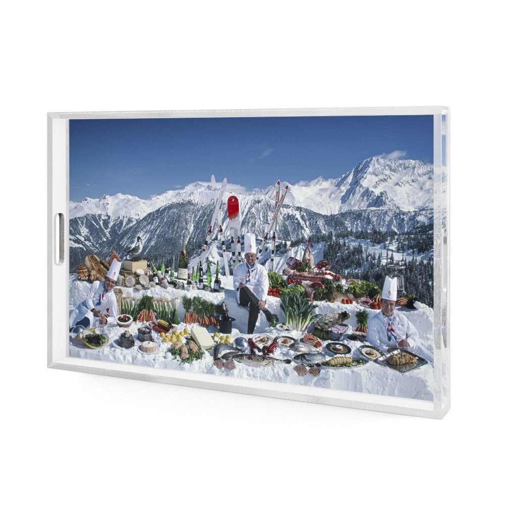 A rectangular clear acrylic serving tray with a white frame, featuring a printed winter mountain scene with chefs and a table of food, shown on a plain white background.