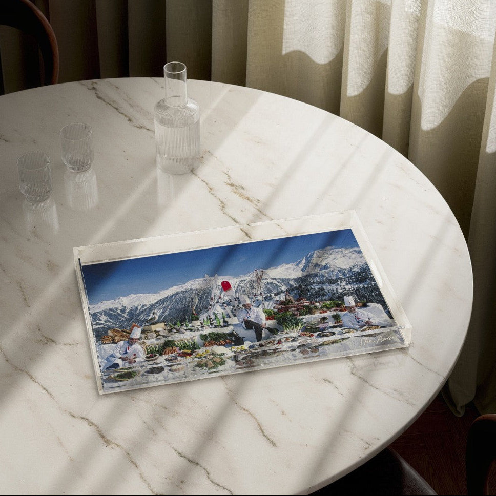 A rectangular printed art print or postcard featuring a snowy mountain village scene, placed on a round white marble table with sunlight and shadows cast across it.