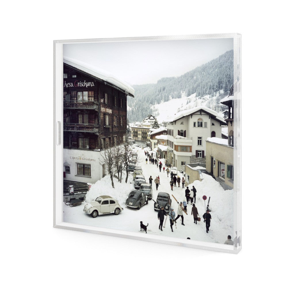 A square framed photographic print depicting a snowy mountain village street with buildings, cars, and small figures of people walking, shown against a plain white background.