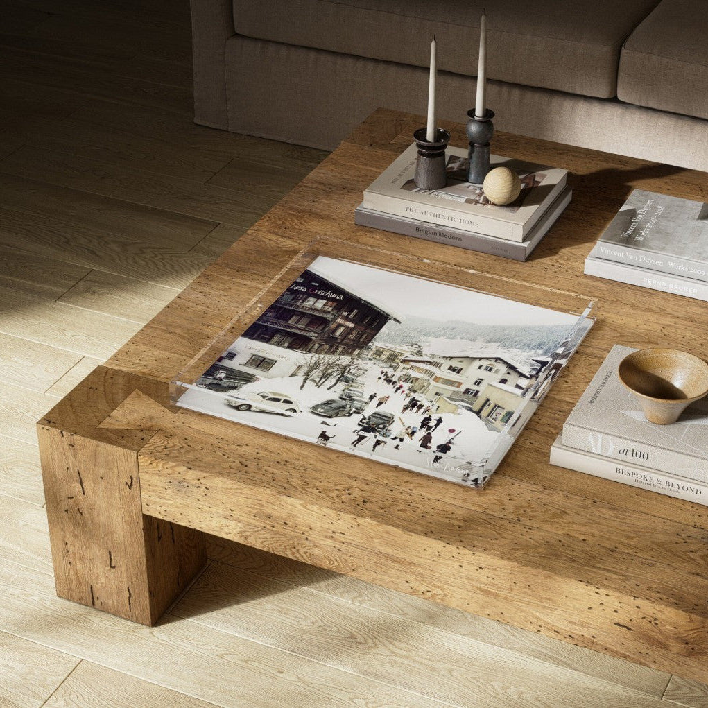 A rustic wooden coffee table in a modern living room setting, styled with books, candles, and a printed art piece on a light wood floor.