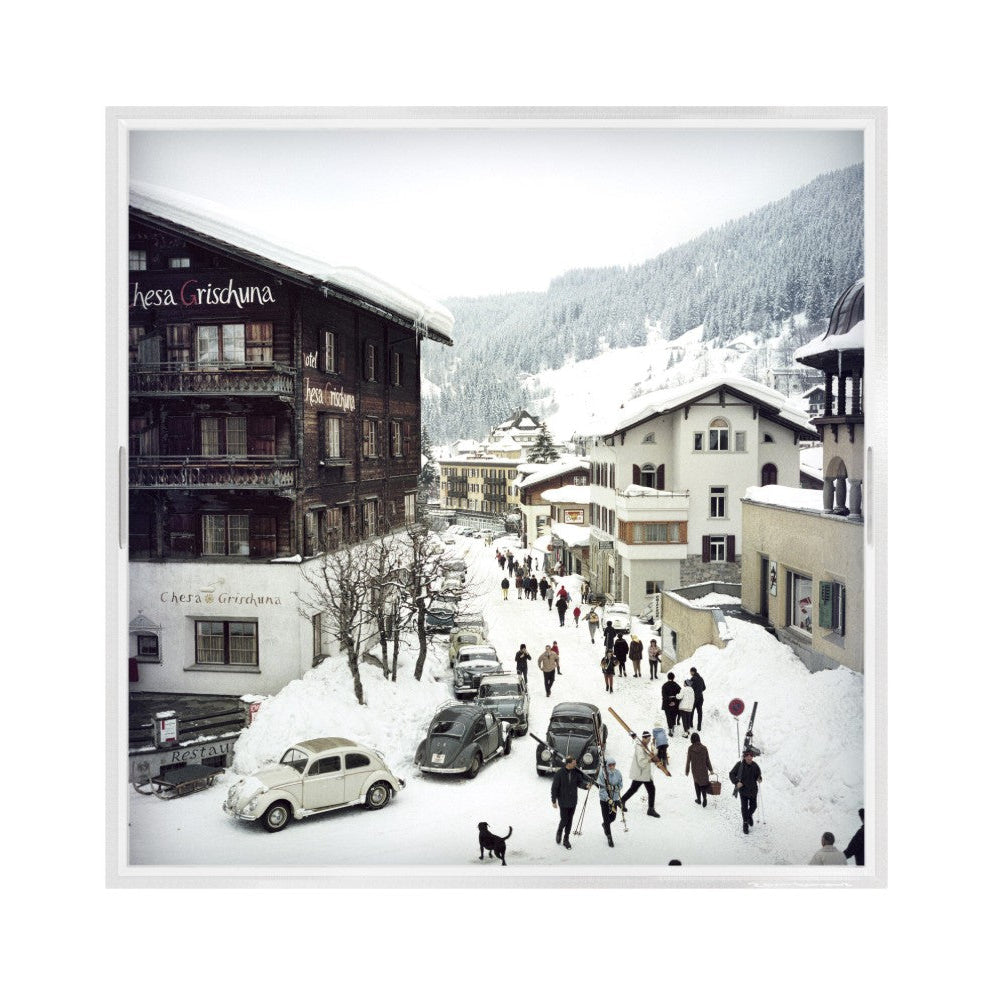 A framed photographic print depicting a snowy mountain village street with alpine buildings, parked cars, and small groups of people walking through deep snow, presented with a white frame.