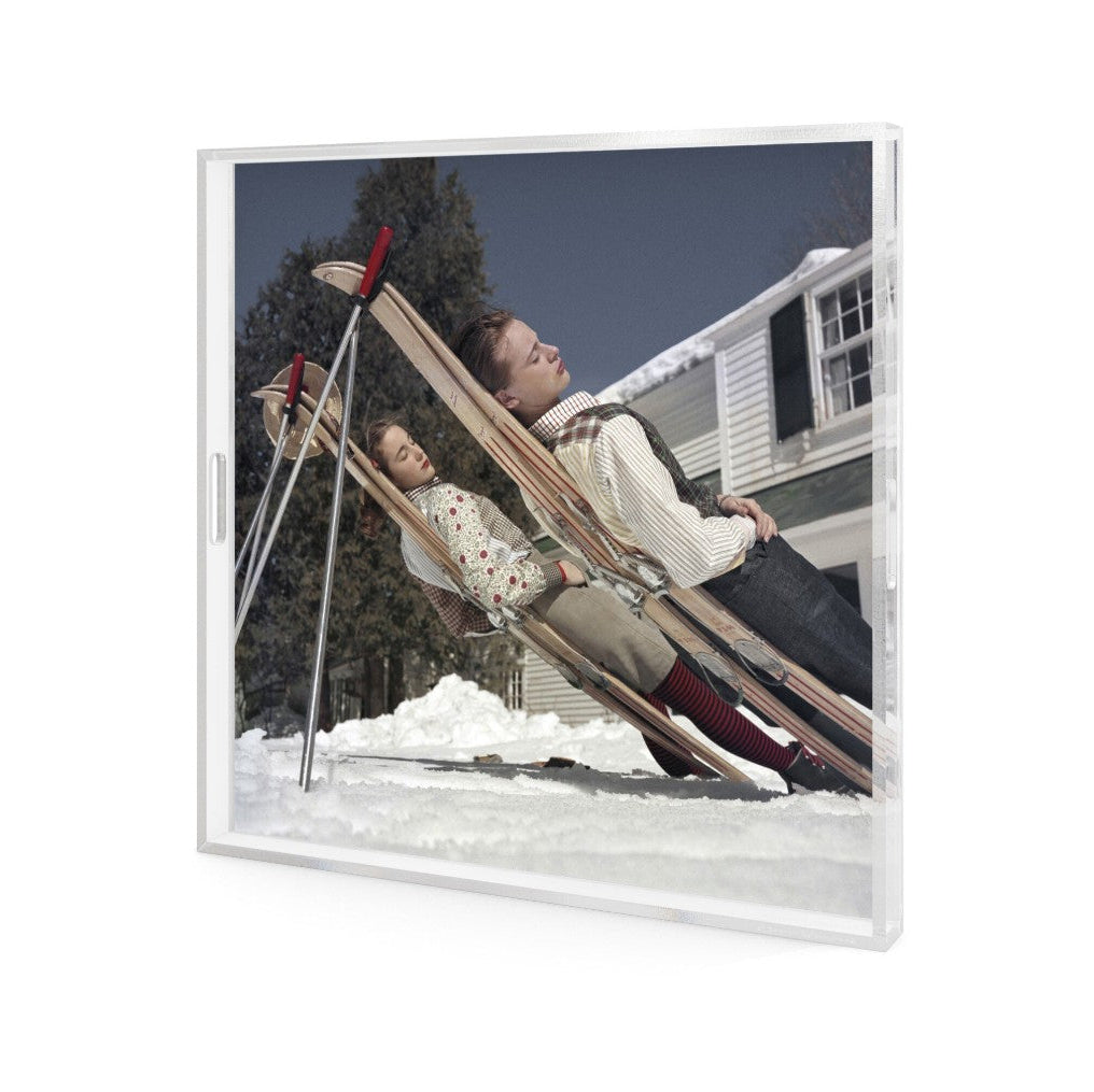 A square framed art print featuring a photograph of a man and child lying on wooden skis in the snow, shown at an angle against a white background.