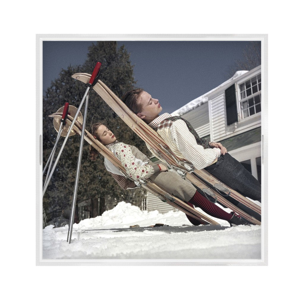 A square framed art print showing a man and a child reclining on wooden sleds in the snow, with a house and evergreen trees in the background.