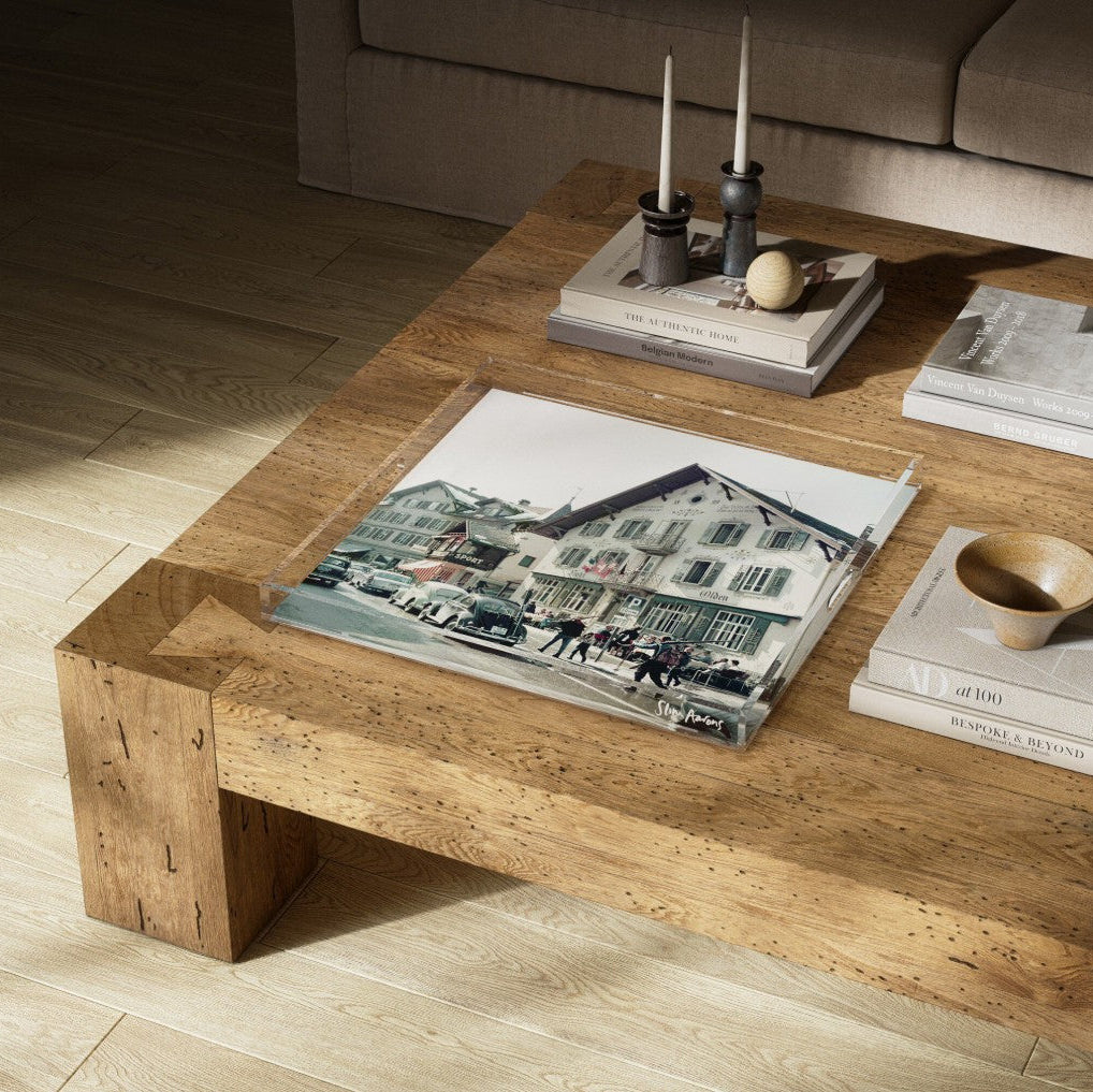 A rustic wooden coffee table in a modern living room setting, styled with books, candles, and a framed print of a European street scene.