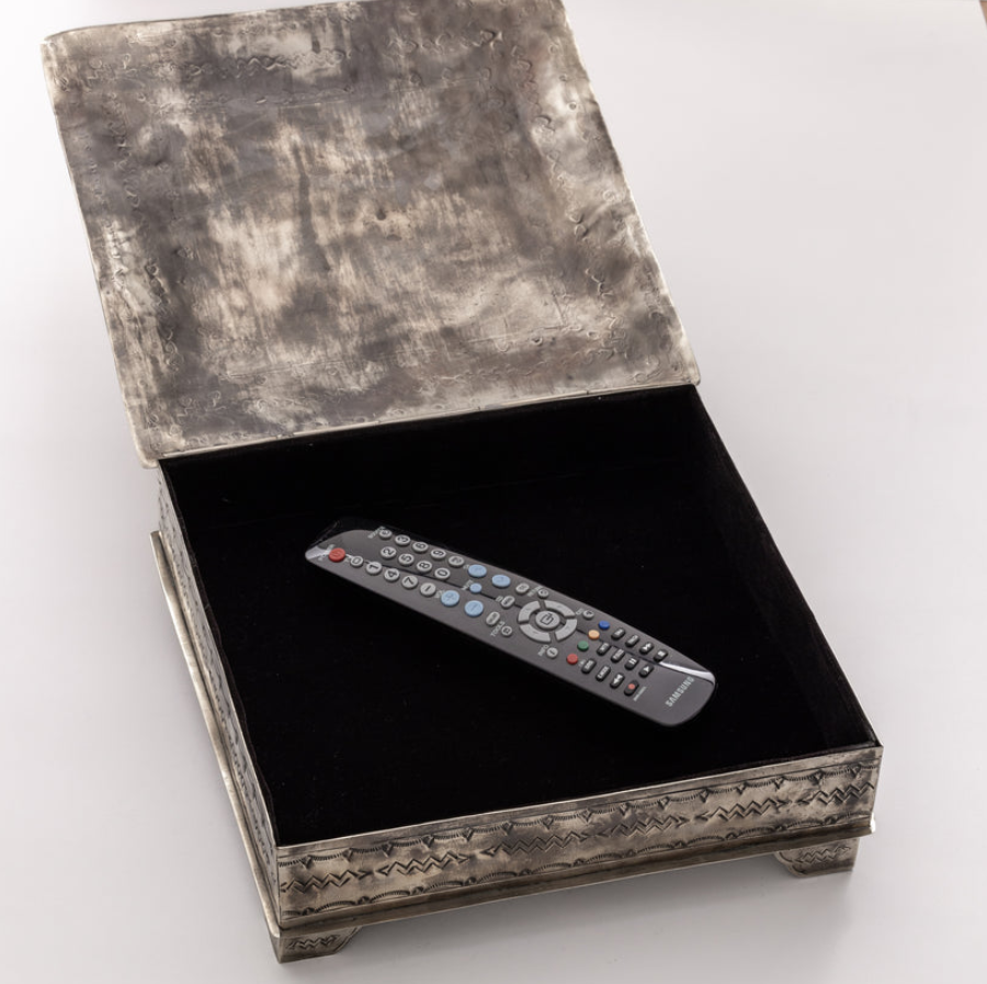 An ornate rectangular silver-toned metal box with a textured finish, shown open on a white background. The interior is lined with black velvet, and a gray remote control rests inside.