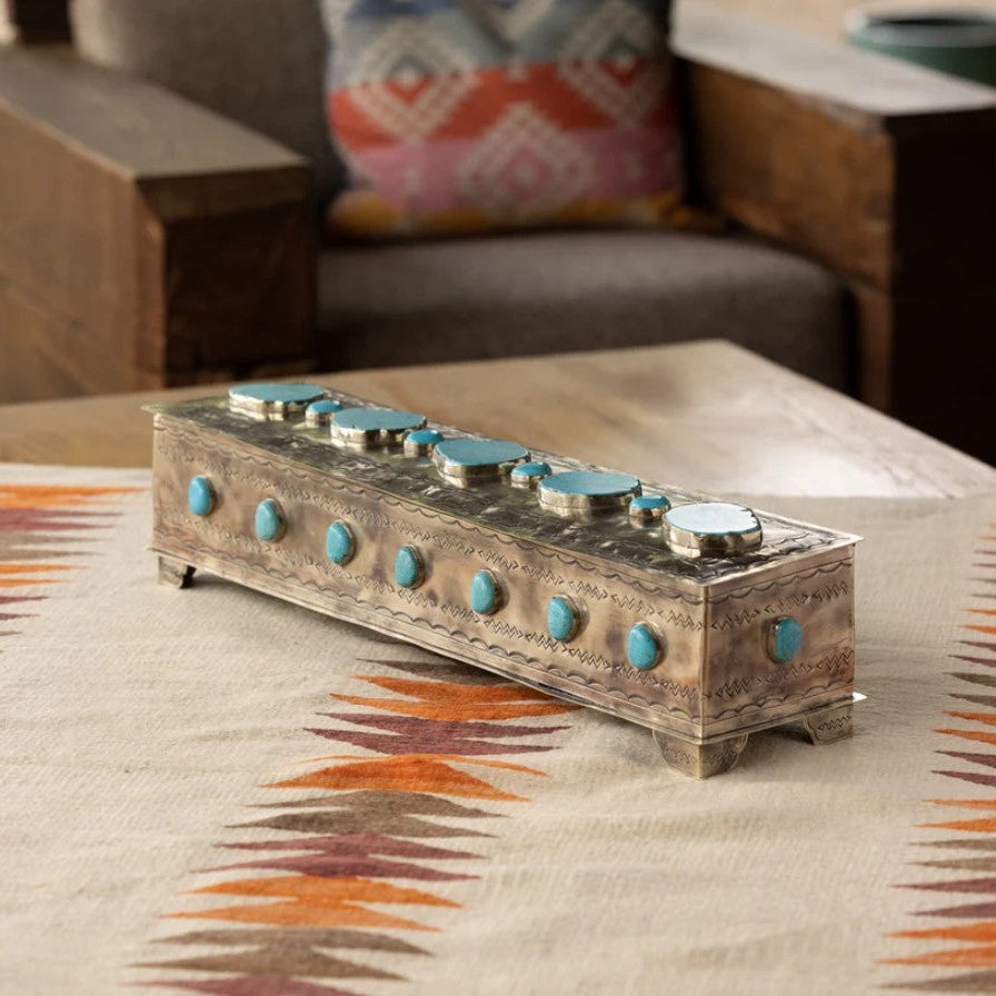 A rectangular silver-toned decorative box with turquoise stone accents, placed on a patterned tablecloth in a living room setting.