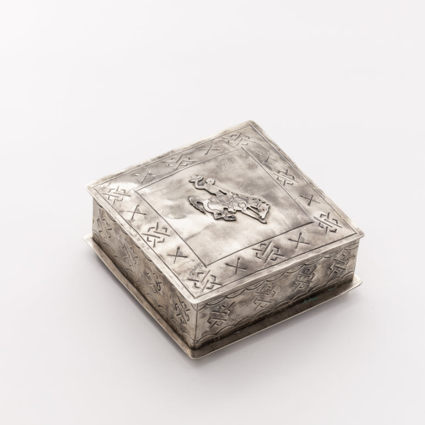 An ornate square silver box with embossed floral and geometric patterns on the lid and sides, featuring a small raised figure detail on the top, photographed on a clean white background.
