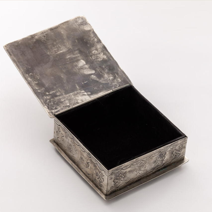 An antique silver-toned jewelry box with an open lid, featuring embossed floral patterns on the sides and a black velvet-lined interior, set against a plain white background.