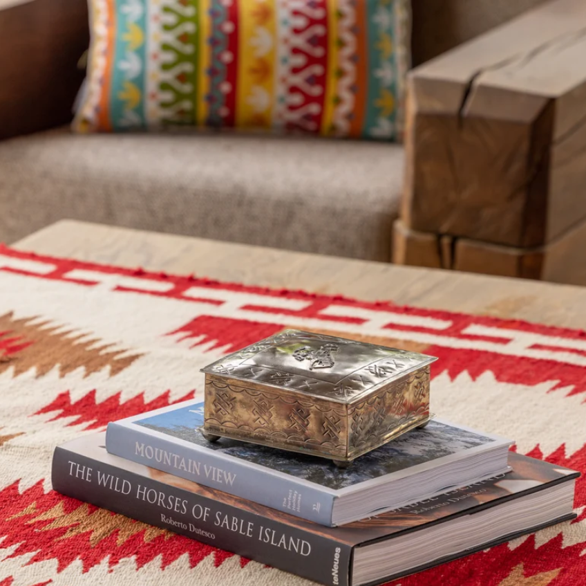 A small ornate silver keepsake box with a carved lid sits on top of two stacked books on a patterned red, white, and beige rug in a living room setting.