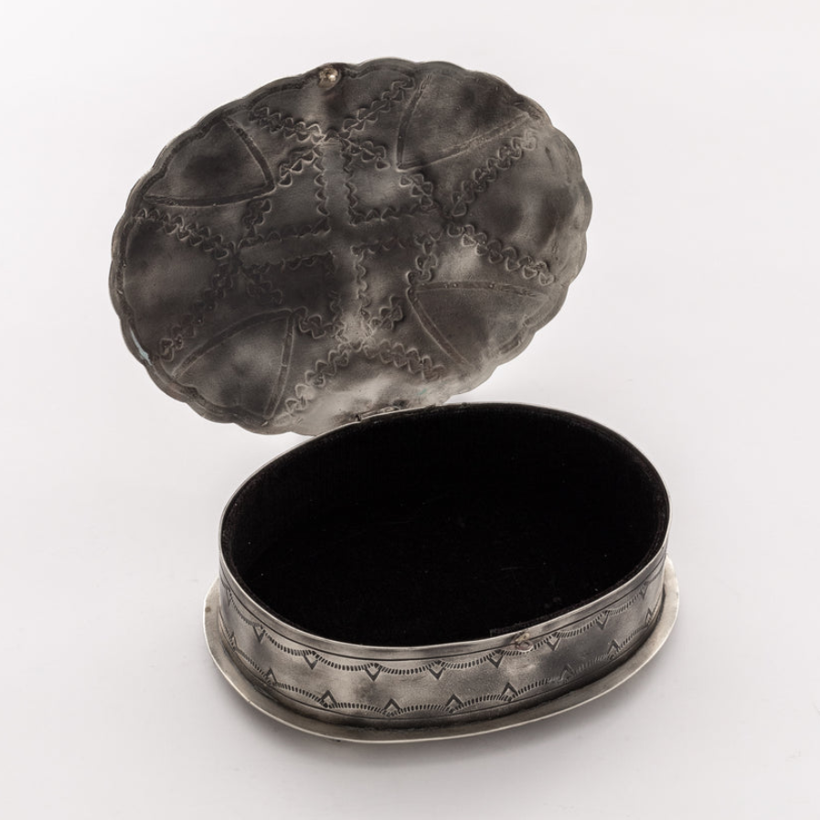 An antique oval silver jewelry box with an open lid, featuring embossed decorative patterns on the lid and a black velvet-lined interior, set against a plain white background.