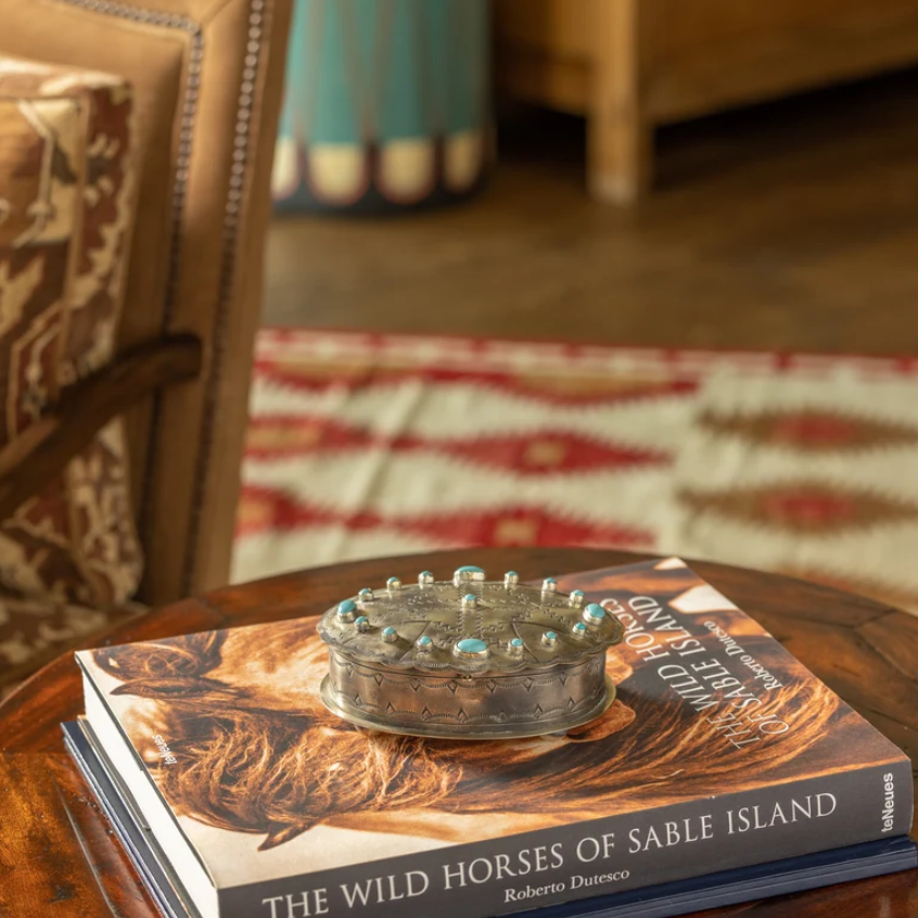 A silver-toned metal keepsake box with turquoise blue stones sits on top of a book titled 'The Wild Horses of Sable Island' on a round wooden table in a cozy, softly blurred living room setting.