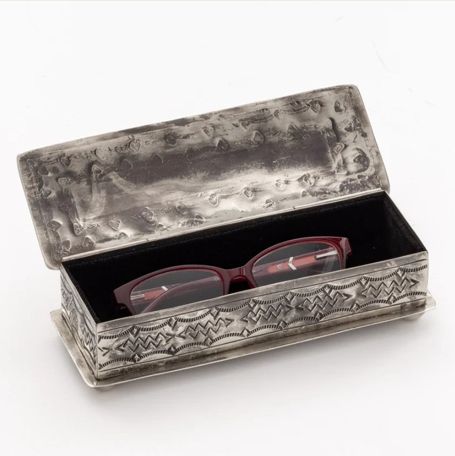 An ornate silver-toned metal box with embossed floral and geometric patterns, shown open with a pair of burgundy eyeglasses inside on a white background.