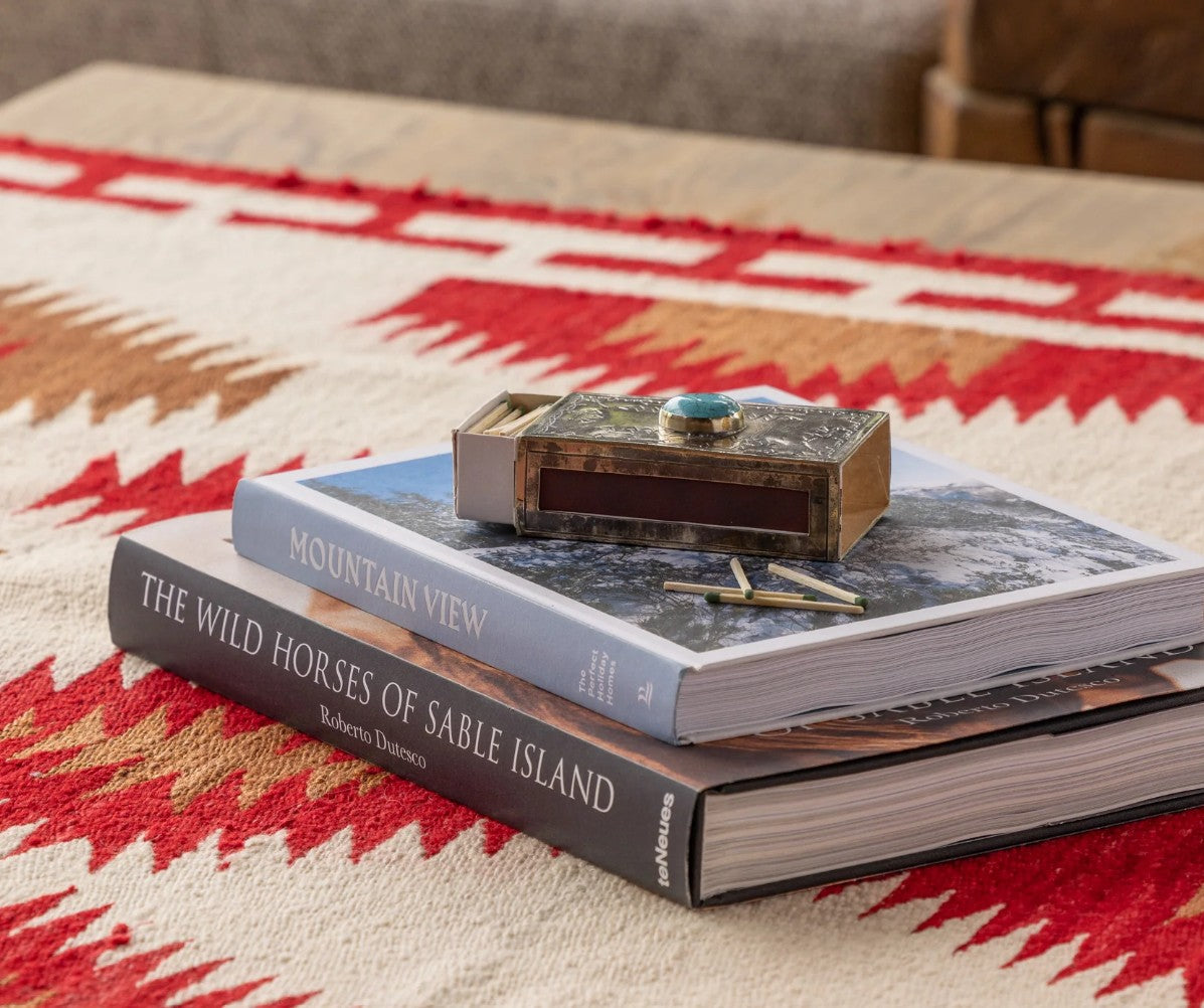 A stack of two books on a patterned rug, with a small decorative box and matchbox on top. The book underneath is titled 'The Wild Horses of Sable Island' by Robert L.