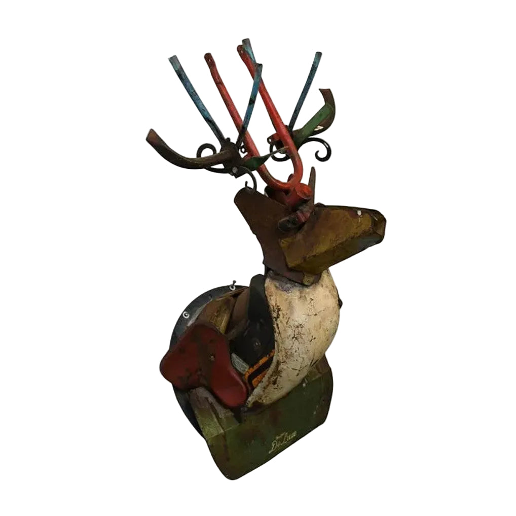 A rustic, handmade metal reindeer sculpture mounted on a circular base, featuring a weathered white body, brown head, red antlers, and green and blue accents, shown against a plain white background.