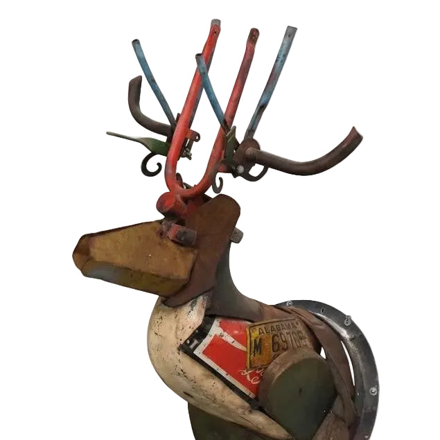 A rustic metal deer sculpture made from salvaged car parts, featuring a weathered body with attached license plates and red-painted antlers, shown against a plain white background.