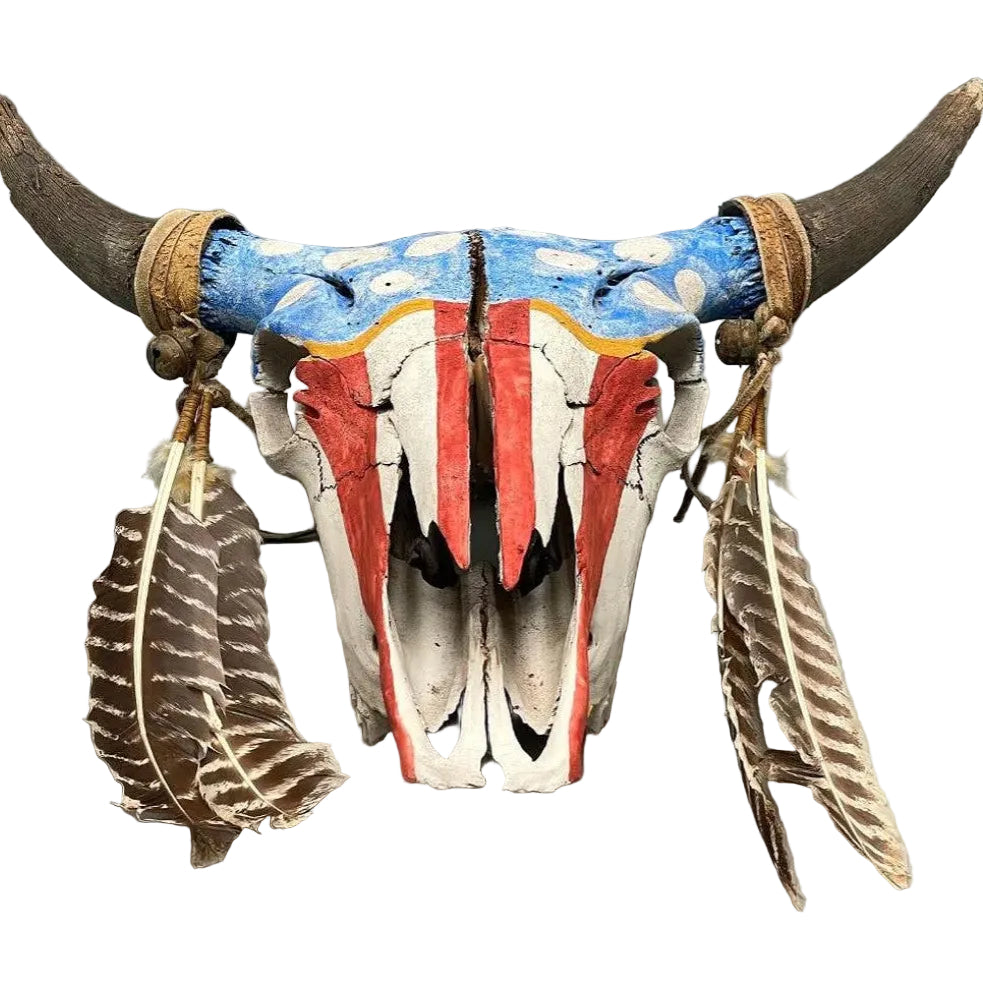 A decorative animal skull with curved horns, painted in red, white, and blue with feather ornaments hanging from the sides, shown against a plain gray background.