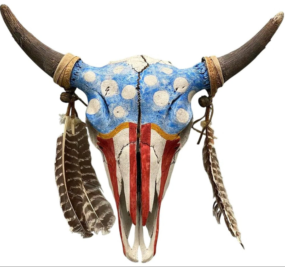 A painted horned skull wall hanging with curved dark brown horns, a blue top panel with white dots, and an American flag-inspired red, white, and blue painted face, adorned with hanging feathers on a white background.