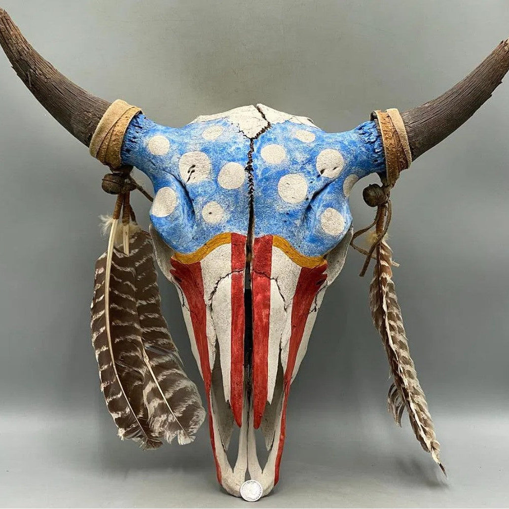 A decorative painted animal skull with curved horns, featuring an American flag-inspired design in blue, white, and red, with hanging feathers and tan wrap accents, photographed against a plain gray studio background.