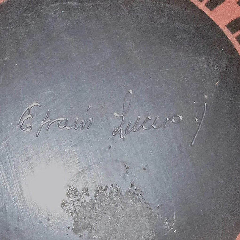 Close-up of the inside of a dark gray ceramic plate or dish, showing the embossed cursive signature 'E. Trués Lucio' and a partial terracotta-colored patterned rim at the edge.