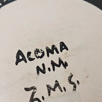 Close-up of the bottom of a white ceramic plate or bowl with black painted decorative edges and handwritten black text reading 'ACOMA N.M' and 'Z.