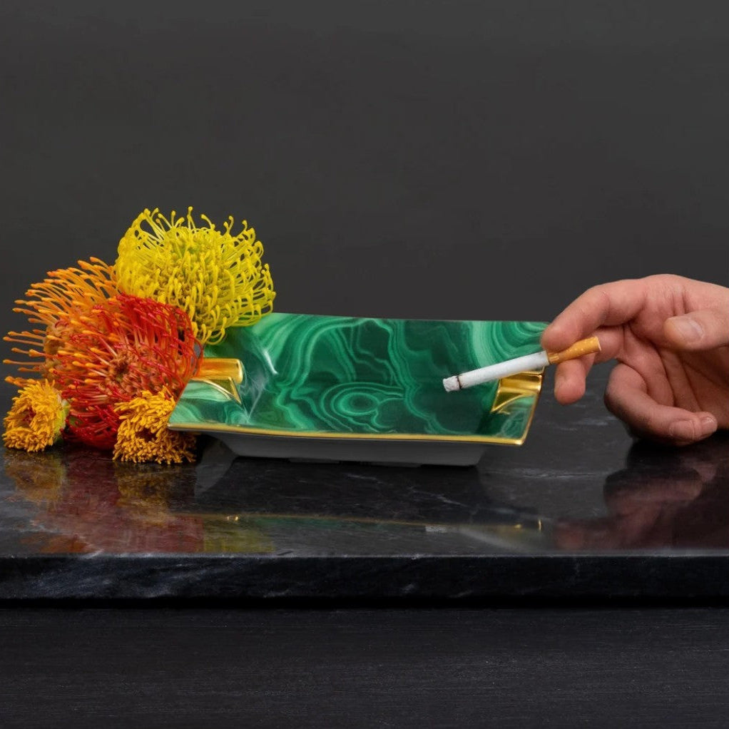 A rectangular green and white marbled ceramic ashtray with gold trim, shown on a dark marble surface with decorative orange and yellow flowers and a hand holding a cigarette.