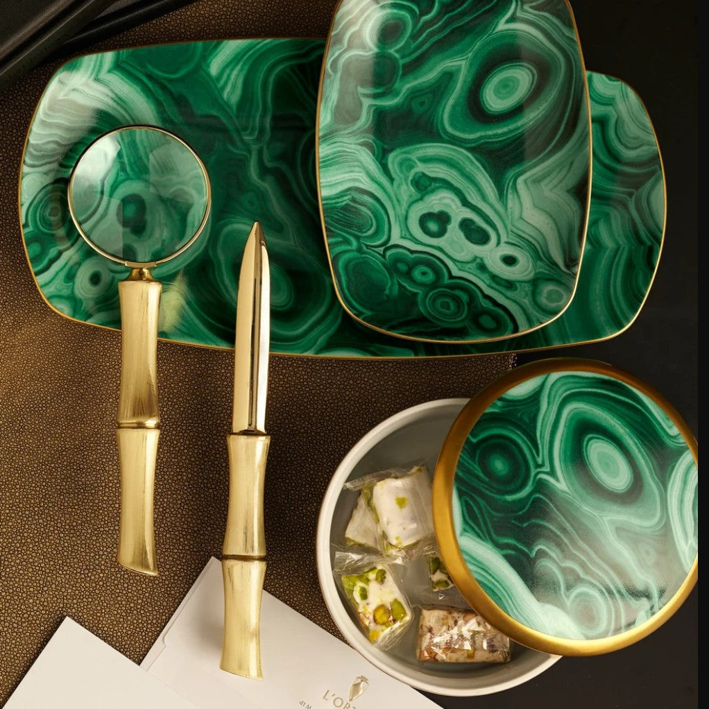 A luxury tableware set featuring a green malachite-patterned ceramic dinnerware collection with gold trim, accompanied by gold bamboo-style utensils and a small bowl of sweets, arranged on a dark textured surface.
