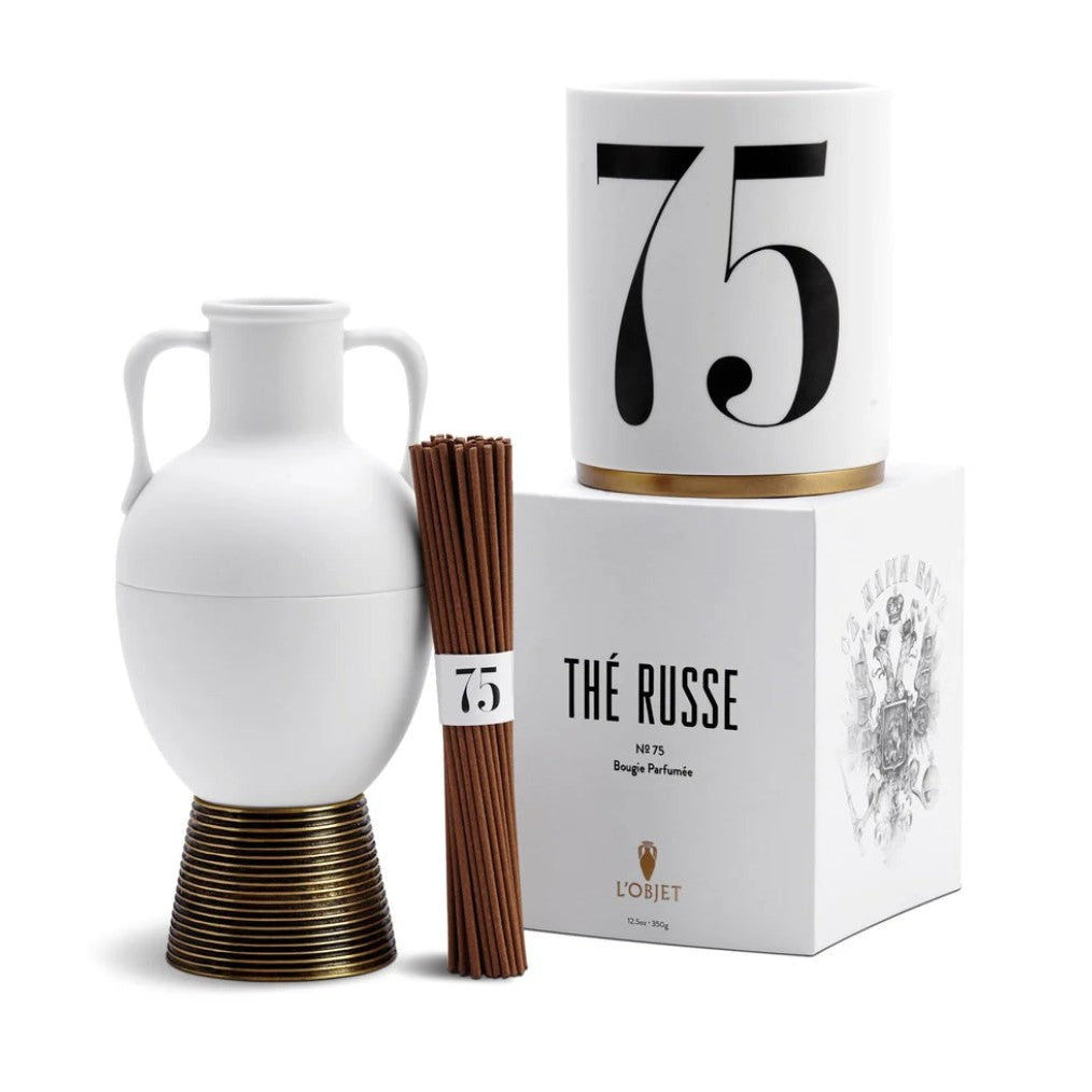 A luxury gift set featuring a white ceramic double-handled vase on a bronze base, a white cylindrical candle with a large black '75' and a gold band, a bundle of incense sticks, and a white branded box reading 'Thé Russe' by L'Orchid.
