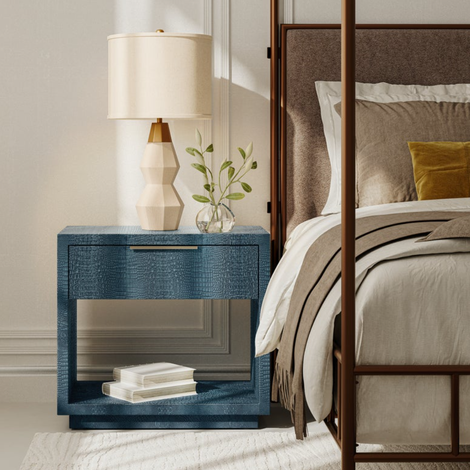 A blue textured nightstand with a mirrored front panel in a styled bedroom setting, next to a bed with neutral-toned bedding and a cream-colored table lamp on top.