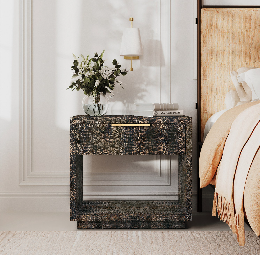 A modern nightstand with a dark textured crocodile-patterned shell finish, featuring a single drawer with a gold handle and an open lower shelf. It is styled in a bedroom setting next to a bed with a beige headboard, holding a glass vase with white flowers and a stack of books.