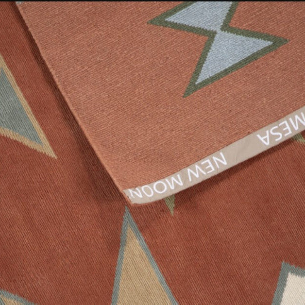 Close-up of a woven rug or textile in a warm terracotta/rust tone with geometric star-like motifs in muted blue-gray, olive green, beige, and cream. A folded edge shows the brand name NEW MOON MESA.