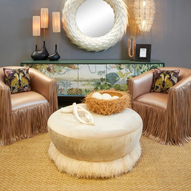 Two fringed armchairs with a round ottoman and decorative elements in a room.