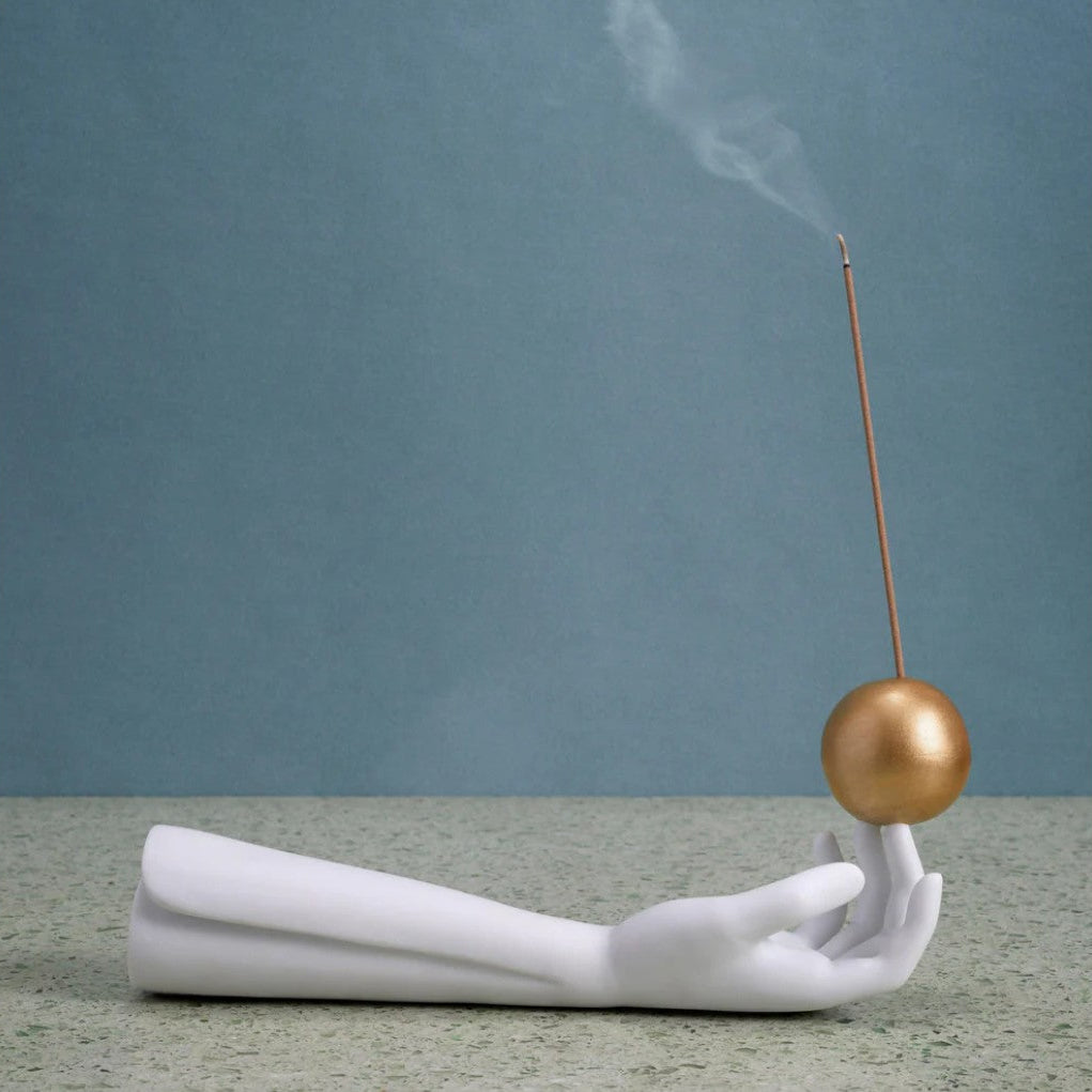 A white sculptural hand holding a metallic gold sphere with a thin rod extending upward, set against a muted blue-gray background.