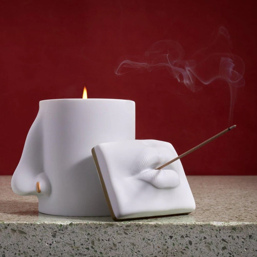 A white ceramic incense holder shaped like a hand holding an incense stick, with a separate white cylindrical candle or vessel behind it and visible smoke rising against a dark red background.
