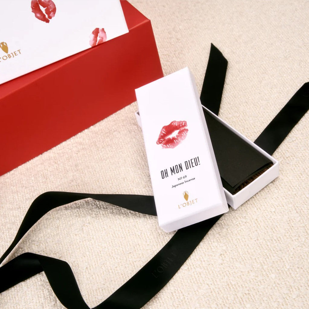 A white rectangular box labeled 'OR MON DIE! L'ORNET' with a red lip print, placed beside an open black-lined box on a beige textured surface. A red L'Ornet box with a white insert and red lip print is partially visible in the background, with a black ribbon draped across the scene.