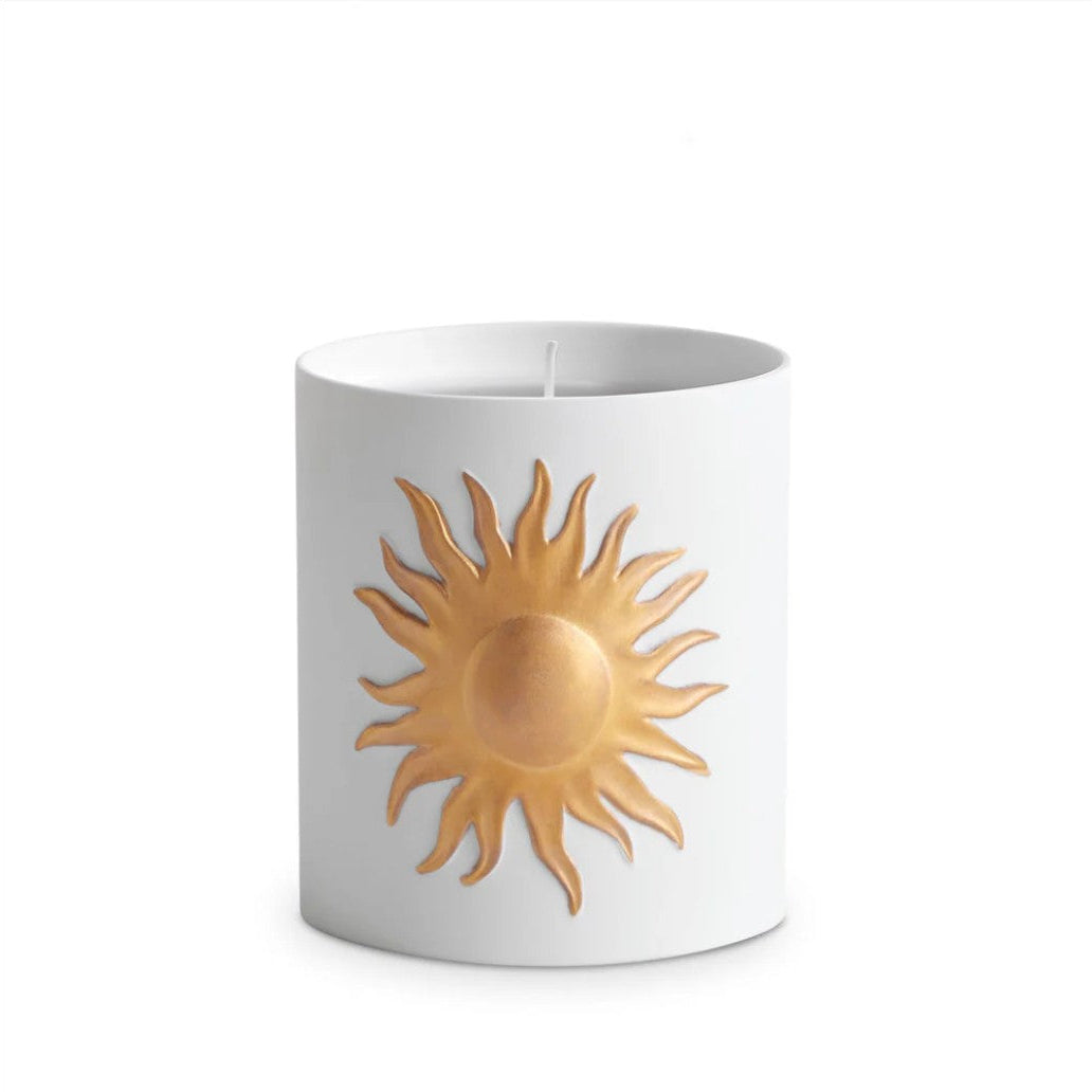 A white cylindrical candle with a raised gold sun emblem on the front, centered against a plain white background.