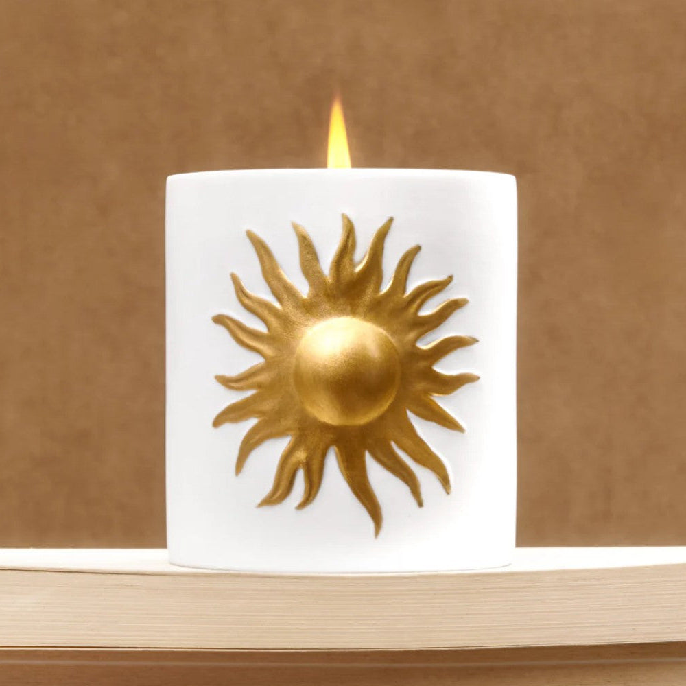 A white cylindrical candle with a lit flame and a raised gold sun emblem on the front, displayed on a light wood surface against a warm brown background.