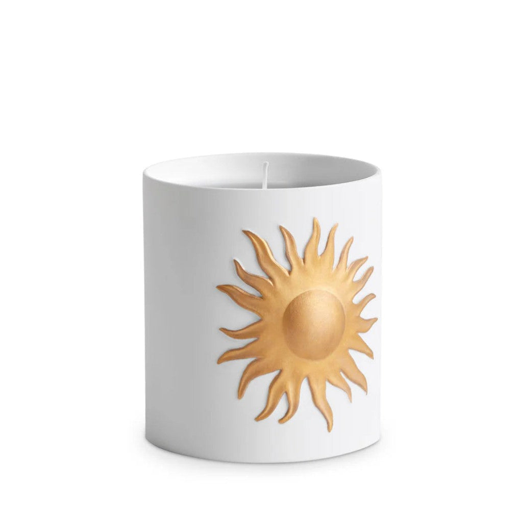 A white cylindrical candle with a metallic gold sun emblem on the front, set against a plain white background.