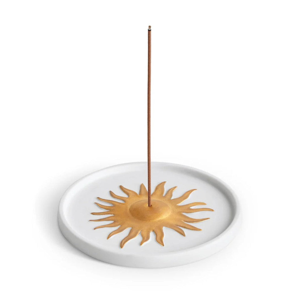 A white ceramic incense holder with a gold sun design in the center, with a single incense stick standing vertically inside it, set against a plain white background.