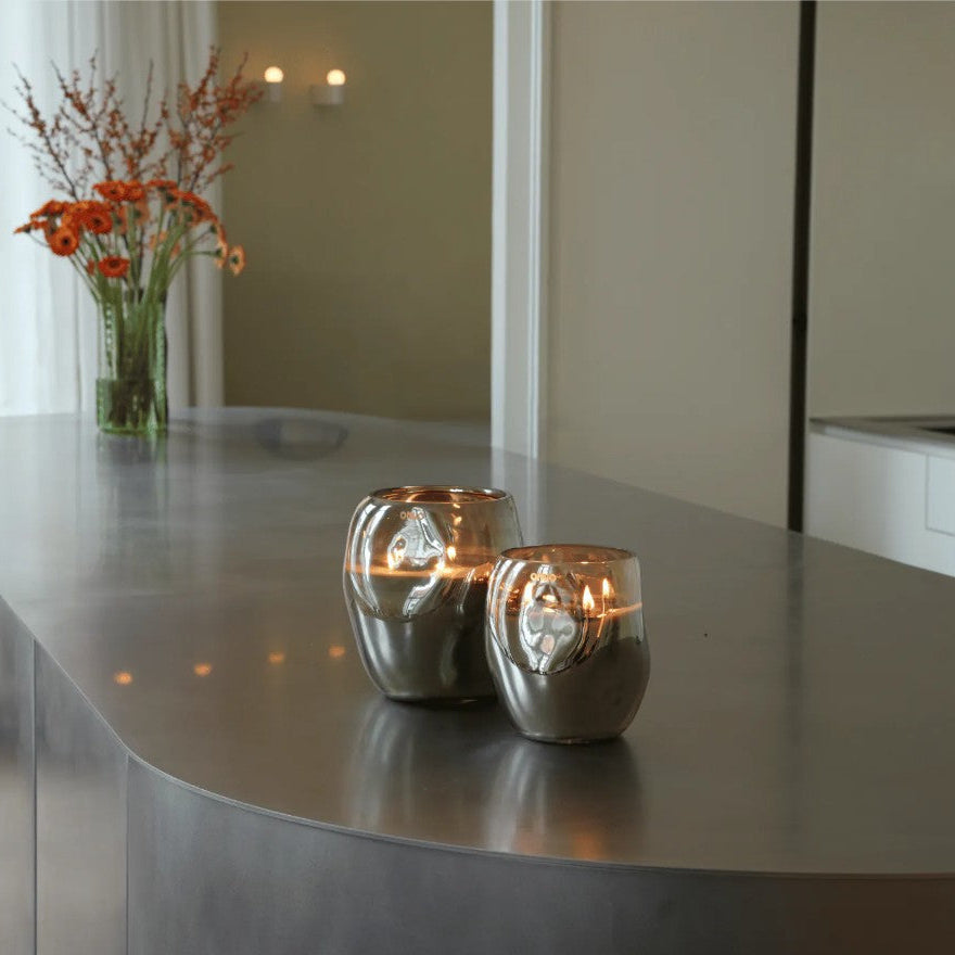 Two small metallic silver glass candle holders with lit candles on a curved gray countertop in a modern kitchen setting.