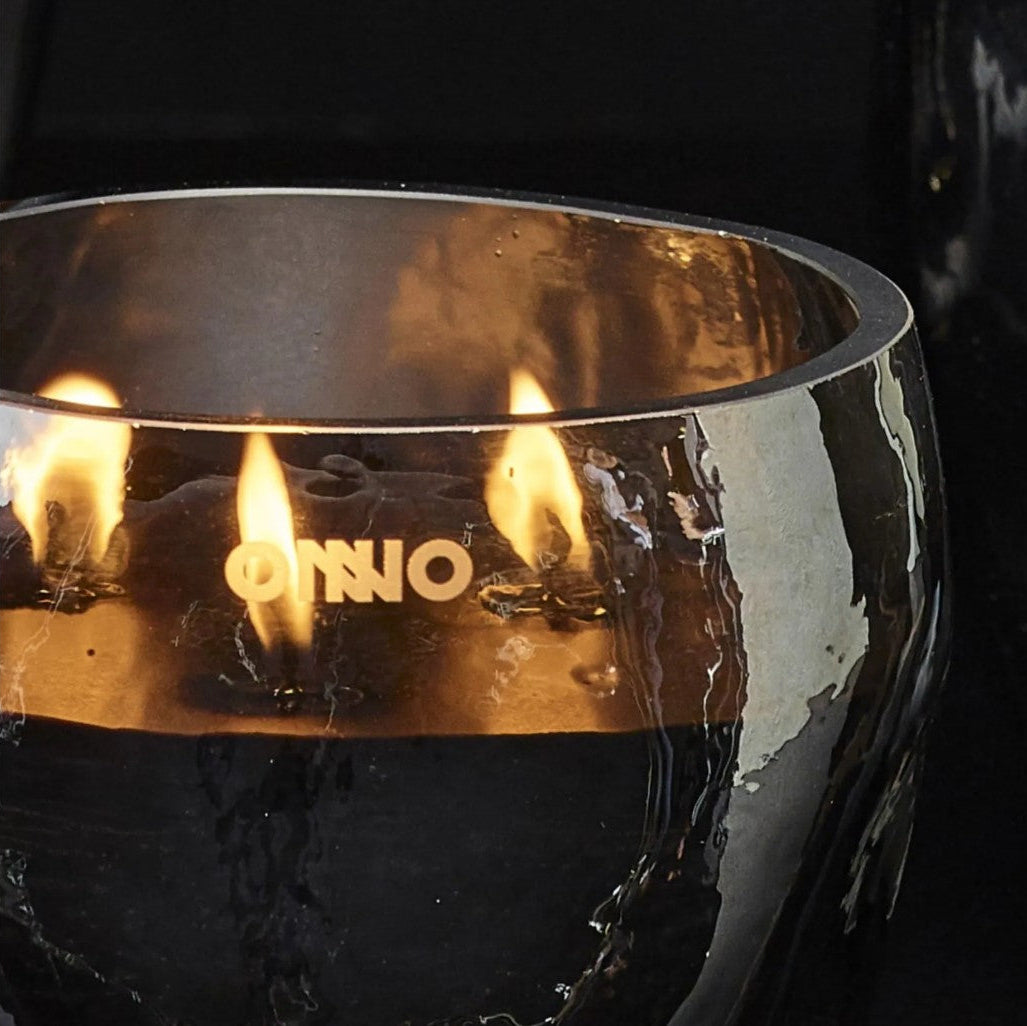 A close-up of a metallic bowl-shaped candle holder with three lit flames and the brand name 'ONNO' printed on the front, set against a dark background.
