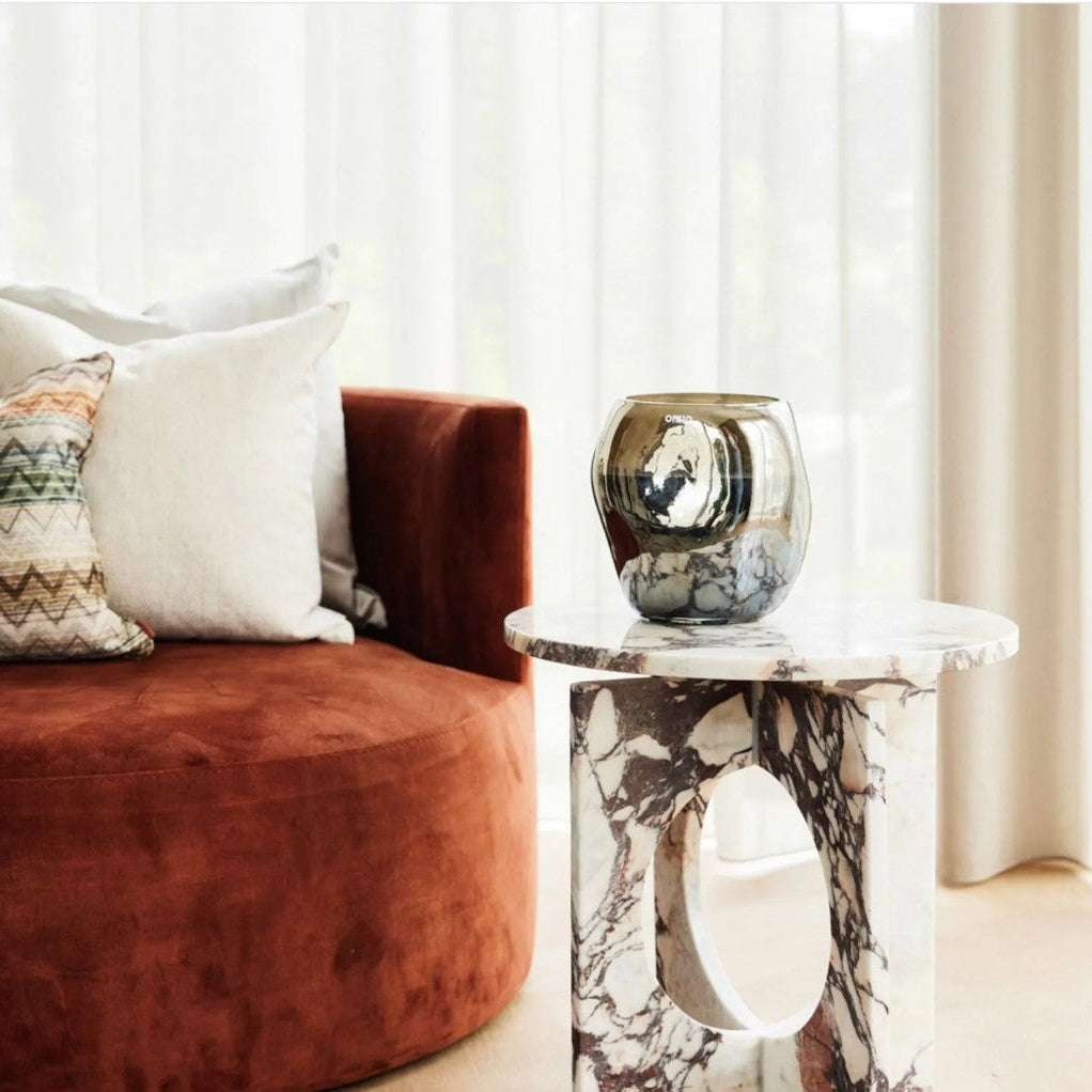 A modern round side table with a white and gray marble pattern and a cutout base, holding a reflective metallic vase, next to a rust-brown velvet sofa in a bright room.