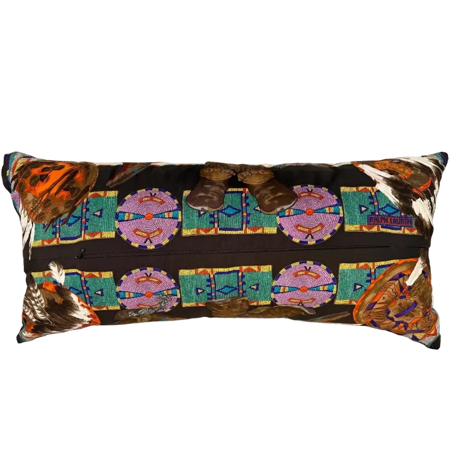 A rectangular lumbar throw pillow with a Southwestern-style pattern featuring turquoise, purple, orange, and brown geometric designs on a black background, resting on a gray textured sofa.