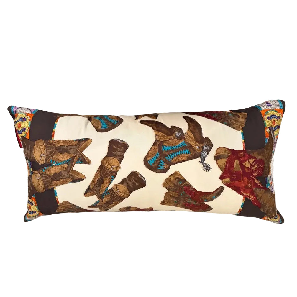 A rectangular lumbar throw pillow featuring a vintage-style print of cowboy boots in brown, red, and turquoise on an off-white background, resting on a gray sofa.